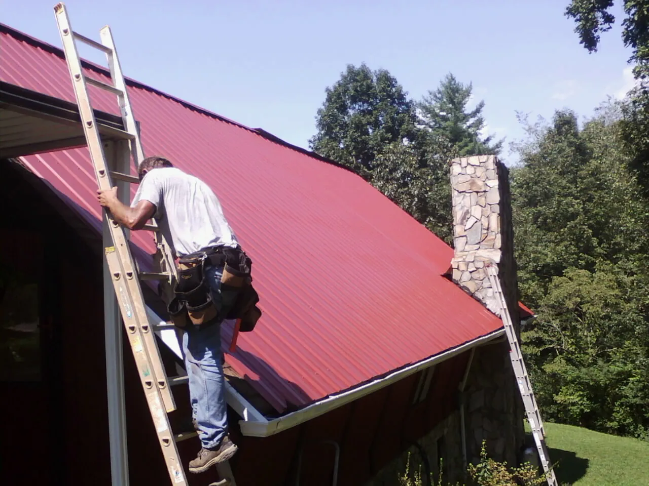 Roofer on a ladder beside a red metal roof and stone chimney — Soffit & Fascia Repair by Roofing Brothers in Hardin